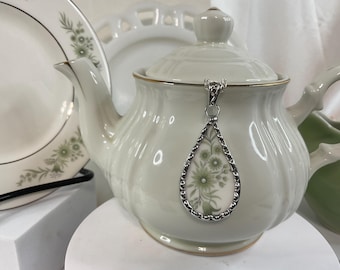 May include: A white porcelain teapot with a silver teardrop pendant featuring a floral design. The pendant hangs from the teapot's lid.