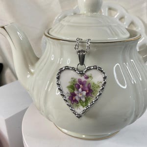 May include: A heart-shaped pendant featuring a floral design of purple and white violets on a white ceramic background, encased in a scalloped silver-toned metal frame and suspended from a silver-toned chain, resting against a white teapot.