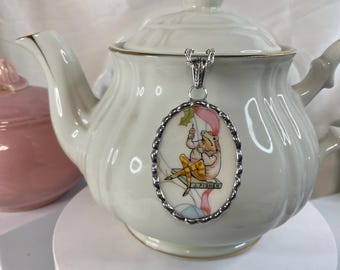 May include: An oval pendant featuring a colorful illustration of a frog wearing yellow striped pants and a jacket, sitting on a book labeled J. FISHER, holding a small umbrella. The pendant has a scalloped silver-tone metal frame and a silver bail.