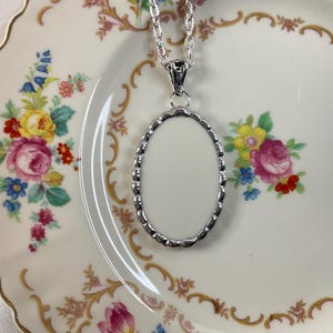 May include: A silver-toned oval pendant necklace featuring a scalloped edge frame with a smooth, blank white center insert. The pendant hangs from a matching silver-toned chain, showcasing a clean, polished metallic finish on the outer border.