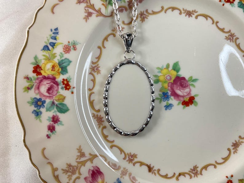 May include: A silver-toned oval pendant necklace featuring a scalloped edge frame with a smooth, blank white center insert. The pendant hangs from a matching silver-toned chain, showcasing a clean, polished metallic finish on the outer border.