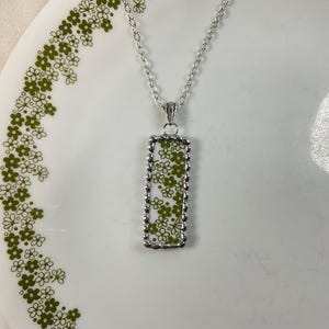 May include: A silver-toned necklace featuring a rectangular pendant with a white background and a repeating olive green floral pattern. The pendant is framed by a scalloped silver edge and hangs from a delicate silver-toned link chain.
