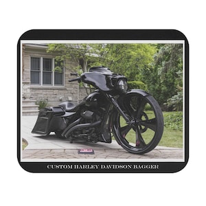 May include: Black custom Harley Davidson Bagger motorcycle with large front wheel and custom paint. The motorcycle is parked on a paved surface in front of a house with a stone facade. The text "CUSTOM HARLEY DAVIDSON BAGGER" is at the bottom.