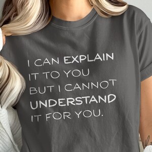 May include: A dark gray t-shirt with the white text: "I CAN EXPLAIN IT TO YOU BUT I CANNOT UNDERSTAND IT FOR YOU." The shirt has short sleeves and a crew neck. The person is wearing a white sweater.
