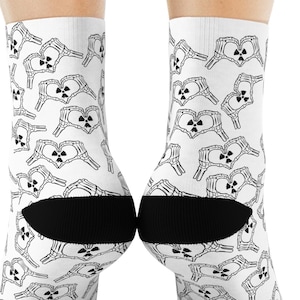 Puede incluir: Calcetines blancos con un patrón en blanco y negro de manos de esqueleto formando corazones, cada uno con un símbolo de radiación. Los calcetines tienen talones y dedos negros.
