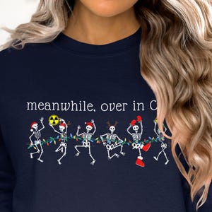 May include: Navy blue sweatshirt with the text "meanwhile, over in C" above a line of dancing skeleton figures. The skeletons are decorated with Christmas lights, Santa hats, and boxing gloves.
