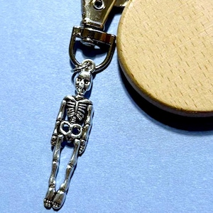 May include: A silver-colored skeleton charm keychain with a wooden round tag. The skeleton charm is detailed and hangs from a silver clasp. The wooden tag is light brown. The background is a light blue.