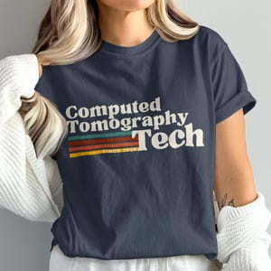 May include: Navy blue t-shirt with the text "Computed Tomography Tech" in white, with a retro-style rainbow stripe design. The text "SINCE 2023" is below the stripes. The shirt is a casual style.