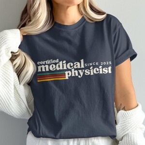 Puede incluir: Camiseta azul marino con el texto "certified medical physicist since 2025" en blanco. El texto tiene un diseño retro con una franja horizontal roja, naranja, amarilla y verde. La camiseta es de cuello redondo.