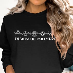 May include: A black crewneck sweatshirt featuring a white graphic print of a heartbeat line interspersed with medical imaging icons including an ultrasound probe, an X-ray symbol, an MRI machine, a radiation sign, and a stethoscope, with the text IMAGING DEPARTMENT.