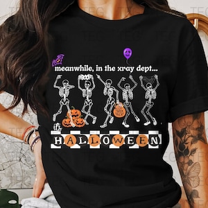 May include: Black t-shirt with a Halloween-themed graphic. The design features dancing skeletons, pumpkins, and the text "meanwhile, in the xray dept... it's HALLOWEEN". A purple bat and balloon are also included in the design.