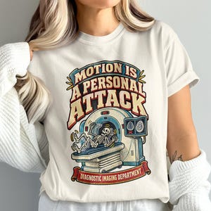 May include: A cream-colored t-shirt featuring a graphic of a skeleton lying on a medical imaging scanner. The text reads "MOTION IS A PERSONAL ATTACK" and "DIAGNOSTIC IMAGING DEPARTMENT" in a vintage-style font with red, blue, and yellow accents.