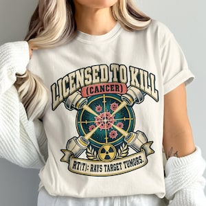May include: A cream-colored t-shirt featuring a graphic design with the text "LICENSED TO KILL (CANCER)" and "RT(T): RAYS TARGET TUMORS." The illustration shows a crosshair targeting a central cell, surrounded by four radiation beams and a radiation symbol.