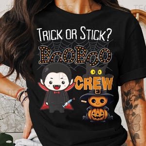 Puede incluir: Camiseta negra con un diseño temático de Halloween. El diseño presenta el texto "Trick or Stick?" encima de "BooBoo Crew". Las ilustraciones incluyen un vampiro, un gato negro y una calabaza con sombrero de bruja.
