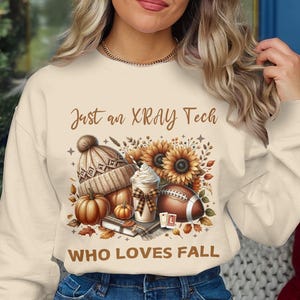 May include: Cream sweatshirt featuring a fall design with a knitted hat, pumpkins, sunflowers, a football, and a coffee drink. Text reads "Just an XRAY Tech WHO LOVES FALL."