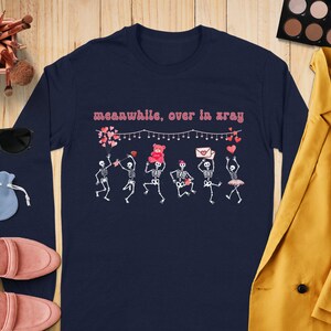 May include: Navy blue long-sleeve shirt with a graphic of dancing skeletons holding hearts, a teddy bear, and an envelope. The text "meanwhile, over in xray" is above the skeletons. A fun, whimsical design.