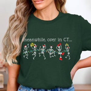 Peut inclure: T-shirt vert foncé avec le texte "meanwhile, over in CT..." au-dessus d'un motif de squelettes dansant décorés de guirlandes lumineuses et d'accessoires. Les squelettes portent des bonnets de Père Noël, tiennent des symboles de radiation et portent des gants de boxe.
