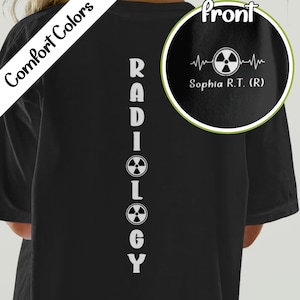 Könnte beinhalten: Schwarzes T-Shirt mit dem Wort "RADIOLOGY" vertikal in Weiß gedruckt. Das Shirt hat auch ein kreisförmiges Design mit einer Herzschlaglinie und einem Strahlungssymbol sowie den Namen "Sophia R.T. (R)". Die Worte "Comfort Colors" sind diagonal gedruckt.