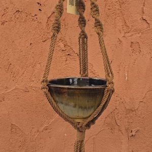 May include: A brown macrame plant hanger featuring twisted cord patterns, light-colored wooden beads, and a central ceramic bowl with a dark, glossy interior and a blended brown and green exterior finish, suspended from a metal ring at the top.