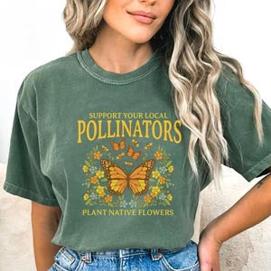 Puede incluir: Camiseta verde con el texto "SUPPORT YOUR LOCAL POLLINATORS" en letras doradas. La camiseta presenta un gráfico de una mariposa monarca con alas naranjas y negras, rodeada de flores y otras mariposas. La frase "PLANT NATIVE FLOWERS" está impresa debajo.