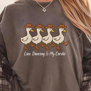 Könnte beinhalten: Dunkelgraues T-Shirt mit einer Grafik von vier Cartoon-Enten mit Cowboyhüten und -stiefeln, die in einer Reihe gehen. Der Text "Line Dancing Is My Cardio" steht darunter. Ein kariertes Hemd ist über den Schultern drapiert.