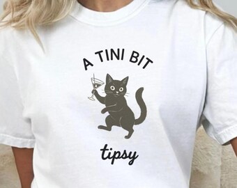 May include: White t-shirt featuring a cartoon cat holding a martini glass. The text above the cat reads "A TINI BIT", and below it, the word "tipsy" is printed in a playful font. The shirt is a casual, everyday wear item.
