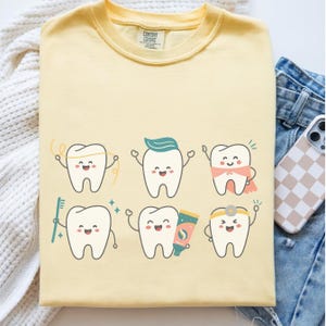 Op de afbeelding: Een lichtgeel Comfort Colors T-shirt met een afbeelding van zes lachende cartoon tanden. Elke tand heeft een ander accessoire, waaronder floss, tandpasta, een tandenborstel en een cape. Het shirt is opgevouwen.
