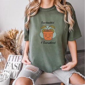 May include: An olive green short-sleeve t-shirt featuring a graphic of a sad, drooping potted plant with a blue teardrop. The text above the plant reads Overstimulated and the text below reads Overwatered in a dark, simple, sans-serif font.