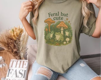 May include: Olive green t-shirt with the text "Feral but cute" above a graphic of three green frogs, mushrooms, and flowers. The shirt is a comfortable, casual style.