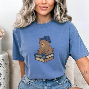 May include: A blue t-shirt featuring a cartoon bear wearing a blue beanie, sitting on top of two books. The books have the word "BERKELEY" printed on them. The shirt is a solid blue color.