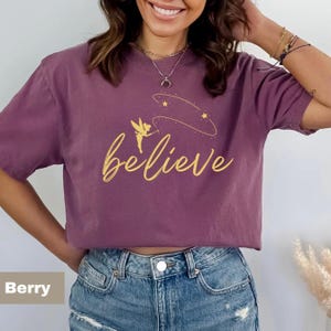 Puede incluir: Camiseta color baya con la palabra "believe" en dorado. Un diseño de hada y estrellas está encima de la palabra. La camiseta es de manga corta y cuello redondo.