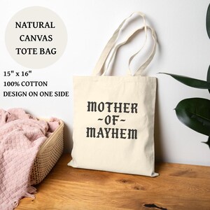 May include: A natural canvas tote bag with the text "MOTHER -OF- MAYHEM" printed in black. The bag is 15" x 16" and made of 100% cotton. The design is on one side. The bag has cream-colored handles.