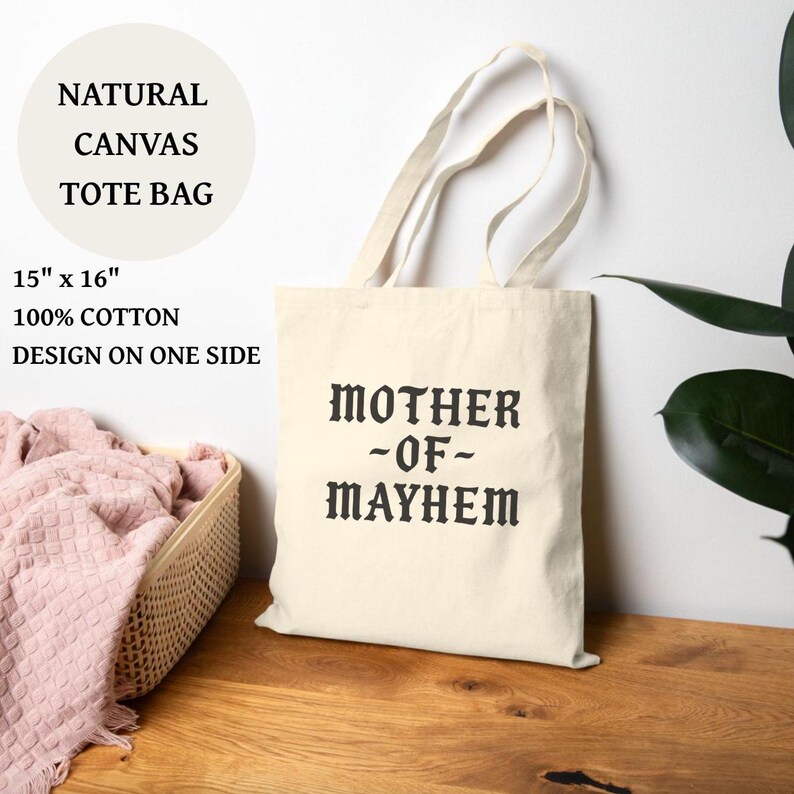 May include: A natural canvas tote bag with the text "MOTHER -OF- MAYHEM" printed in black. The bag is 15" x 16" and made of 100% cotton. The design is on one side. The bag has cream-colored handles.