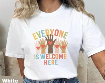 May include: White t-shirt with the text "EVERYONE IS WELCOME HERE" in colourful letters. The design features hands of various skin tones, each with a heart in the centre, symbolising inclusivity and acceptance.