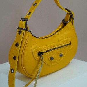 May include: A small, yellow leather handbag with a curved shape and a top handle. The bag has a zipper closure and a small, dangling strap.
