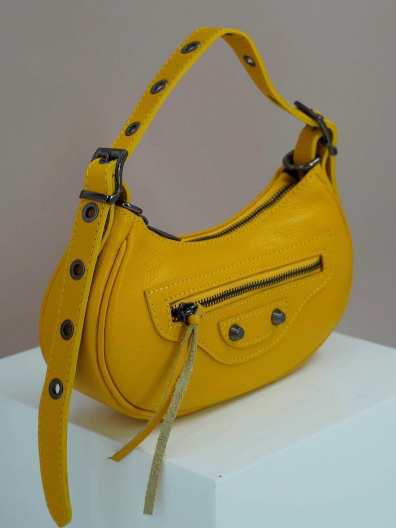 May include: A small, yellow leather handbag with a curved shape and a top handle. The bag has a zipper closure and a small, dangling strap.