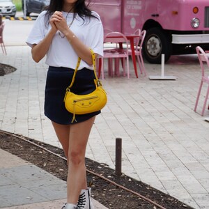 May include: A woman wearing a white t-shirt, a dark blue skirt, and white platform boots. She is carrying a yellow handbag with a strap. The woman is standing in front of a pink ice cream truck.