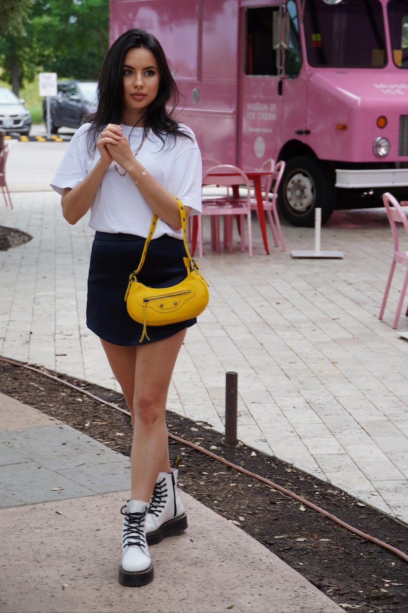 May include: A woman wearing a white t-shirt, a dark blue skirt, and white platform boots. She is carrying a yellow handbag with a strap. The woman is standing in front of a pink ice cream truck.