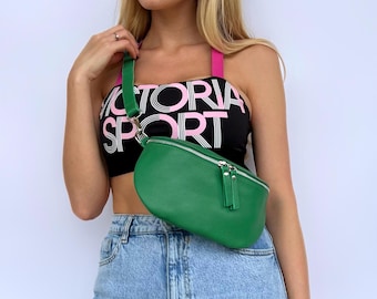 Puede incluir: Una riñonera de cuero verde con una cremallera plateada y una correa negra. La riñonera la lleva puesta una persona que lleva un sujetador deportivo negro y rosa con el texto "VICTORIA SPORT" impreso en él.