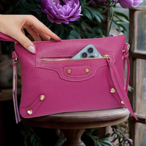 May include: A bright pink leather clutch bag with a gold zipper and a phone in the front pocket. The bag has a gold chain strap and a tassel.