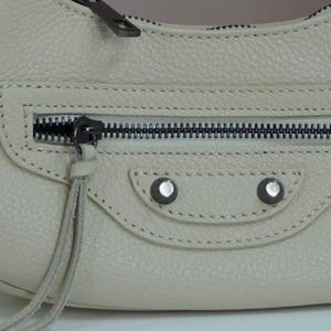 May include: A light gray leather handbag with a zipper closure and silver hardware. The bag has a flap with a stitched detail and two silver studs.
