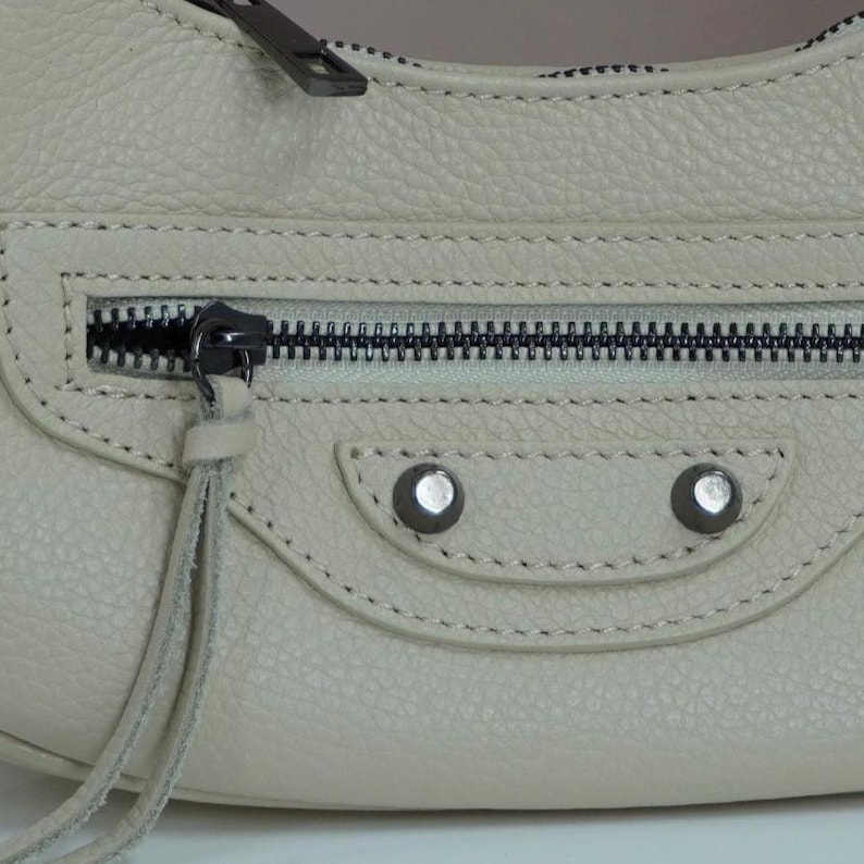 May include: A light gray leather handbag with a zipper closure and silver hardware. The bag has a flap with a stitched detail and two silver studs.
