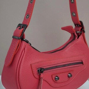 May include: A coral-colored leather handbag with a top zipper closure and a small flap with a tassel. The bag has a short shoulder strap with silver grommets and a buckle.