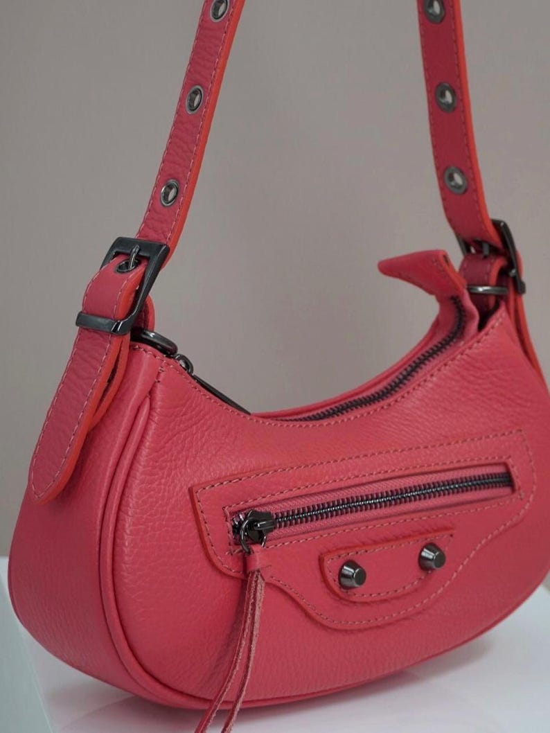 May include: A coral-colored leather handbag with a top zipper closure and a small flap with a tassel. The bag has a short shoulder strap with silver grommets and a buckle.