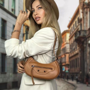 May include: A woman wearing a white blazer and a brown hat is carrying a brown leather handbag with a flap closure and a zipper. The bag has a small tassel hanging from it.