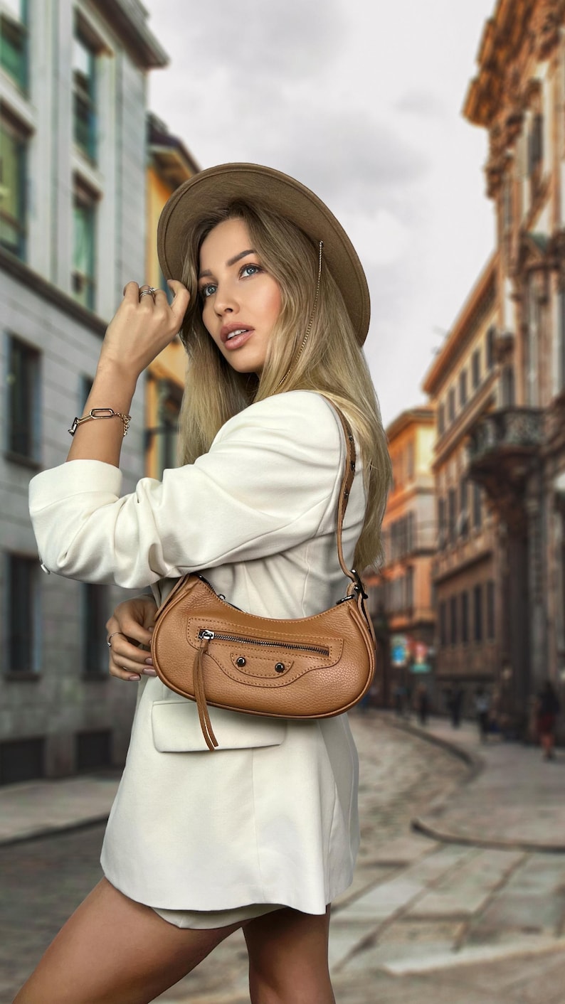 May include: A woman wearing a white blazer and a brown hat is carrying a brown leather handbag with a flap closure and a zipper. The bag has a small tassel hanging from it.