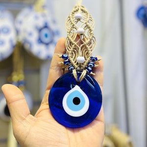 May include: A blue glass Evil Eye amulet with a white and light blue eye design, suspended from a macrame cord. The amulet is held in a hand, with other similar amulets visible in the background.