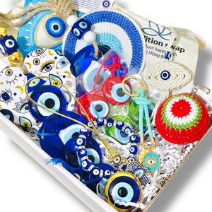 May include: A white gift box filled with various blue and white evil eye themed items. Includes beaded bracelets, keychains, decorative ornaments, and a crocheted red, white, and green ball. The box is lined with white paper and contains a small bag with text.