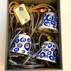 May include: Three white ceramic bells with blue and white evil eye designs are presented in a cardboard box with shredded paper. Each bell hangs from a jute rope with colourful beads. A small tag is attached to the rope.