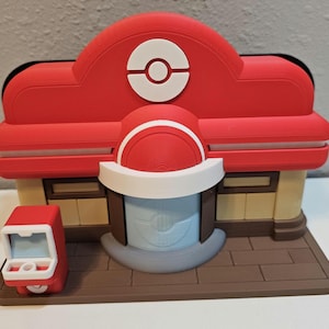 Op de afbeelding: Een 3D-geprint model van een rood en wit gebouw met een cirkelvormig logo bovenop. Het gebouw heeft een bruine basis en een kleine rode machine aan de zijkant. Het gebouw heeft een lichtblauwe deur en ramen.